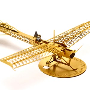 May include: A gold metal model of a 1906 Wright Flyer aeroplane with a pilot figure. The model is on a round base with the inscription "Hubert Latham" and "1906 Wright Flyer" in gold lettering.