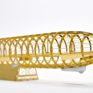 Puede incluir: Un modelo de metal dorado del dirigible Graf Zeppelin. El modelo está detallado con un marco de alambre y el nombre "Graf Zeppelin" es visible en el lateral.