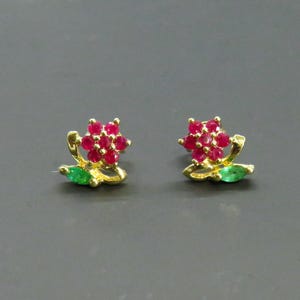 May include: A pair of gold-coloured stud earrings. Each earring features a cluster of small, round, red gemstones, resembling a flower. A green gemstone leaf is at the base of each flower. The earrings are set against a grey background.