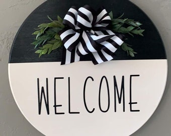 Peut inclure: Une pancarte ronde en bois de bienvenue avec un nœud rayé noir et blanc et de la verdure. La pancarte est peinte en noir sur la moitié supérieure et en blanc sur la moitié inférieure avec le mot "WELCOME" écrit en noir sur la moitié blanche.