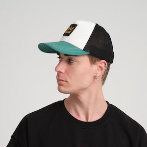 May include: A person wearing a black t-shirt with a small pink logo that says "VELO". The person is also wearing a white and green trucker hat with a black mesh back. The hat has a gold crown logo on the front.