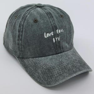 Peut inclure: Une casquette de baseball en denim délavé gris foncé avec un texte brodé blanc qui dit "Love You, Bye".