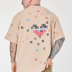 Può includere: Una t-shirt beige a maniche corte con un design a cuore pixelato colorato sul retro. Il cuore è composto da piccoli quadrati di diversi colori e le lettere "YS" in ogni quadrato.