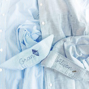 Puede incluir: Una camisa de rayas azul claro y blanco con el nombre Gran bordado y un icono de concha, junto a una camisa azul claro lisa con el nombre Alison bordado y una pequeña ramita en el puño.