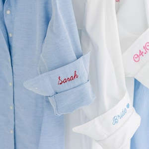 May include: A light blue button-down shirt with the name Sarah embroidered in red on the cuff, alongside white button-down shirts featuring pink embroidery of the date 04.10 and blue embroidery of the word Bride with a small heart icon.