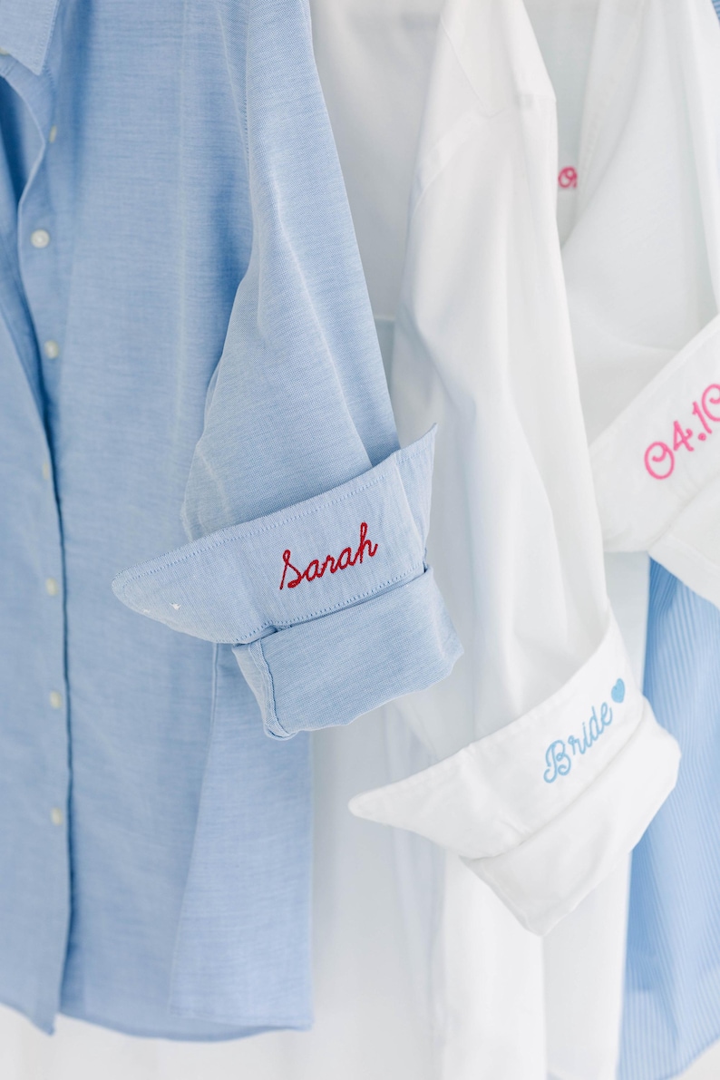 May include: A light blue button-down shirt with the name Sarah embroidered in red on the cuff, alongside white button-down shirts featuring pink embroidery of the date 04.10 and blue embroidery of the word Bride with a small heart icon.