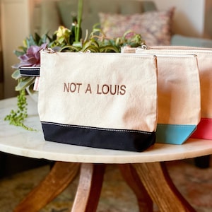 May include: Canvas zipper pouches with the embroidered text "NOT A LOUIS". The pouches have a cream-colored body with a black base. Other pouches have blue and pink bases. The pouches are on a marble table.