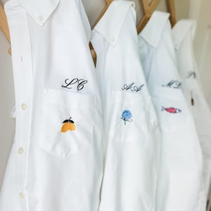 May include: A row of white button-down shirts hanging on a wooden rack. Each shirt features a chest pocket with custom black embroidered initials above a unique colorful design, including yellow lemons, a blue hydrangea, and a small red fish.