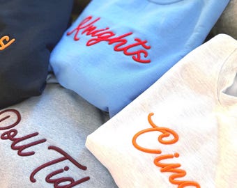 Puede incluir: Una colección de tres sudaderas de cuello redondo en negro, azul claro y gris jaspeado. La sudadera azul claro presenta la palabra Knights bordada en rojo, la gris dice Roll Tide en granate y la blanca muestra Cincy en color naranja.
