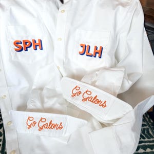 Op de afbeelding: Twee witte overhemden met lange mouwen en knoopsluiting, voorzien van geborduurde details. Het ene overhemd heeft de initialen SPH in oranje en blauw op de zak, het andere JLH. Beide manchetten zijn geborduurd met de oranje tekst Go Gators.