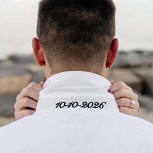 Peut inclure: Chemise blanche à col avec la date "10-10-2026" brodée en fil noir. Le dos de la chemise est visible, avec les mains d'une personne tenant doucement le col. L'arrière-plan est flou, suggérant un cadre extérieur.