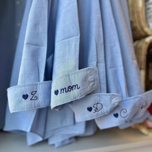 Puede incluir: Camisas celestes con puños bordados. Los puños muestran las palabras "mom" y las letras "Z", "D" y "C", cada una acompañada de un pequeño corazón azul. Las camisas están cuidadosamente dobladas.