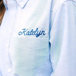 Könnte beinhalten: Ein hellblaues Hemd mit Knopfleiste, spitzem Kragen, langen Ärmeln und einer Brusttasche. Der Name Katelyn ist in dunkelblauer Schreibschrift direkt über der Tasche eingestickt. Das Hemd hat weiße Knöpfe und einen geknoteten Saum vorne.