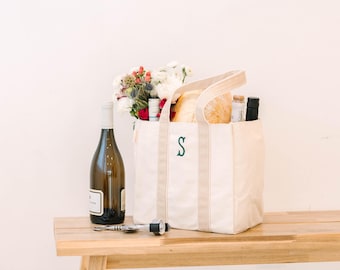 May include: A cream-colored canvas tote bag with beige handles and a dark green embroidered letter S on the front. The bag is filled with a bouquet of white and red flowers and a loaf of bread, accompanied by a wine bottle and a metal corkscrew.