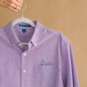 Könnte beinhalten: Ein hellviolettes Langarmhemd mit Knopfleiste, Kragen und einer Brusttasche. Auf der Tasche ist der Name Breta in blauer Schreibschrift eingestickt. Ein Markenetikett von Port Authority ist am inneren Kragen des Hemdes zu sehen.