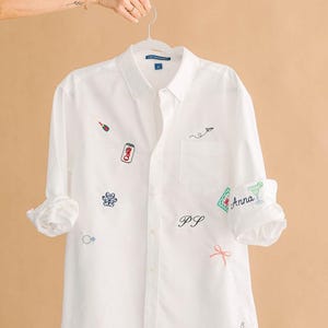 May include: White button-down shirt with embroidered details. Features a champagne bottle, a Coke can, a paw print, a ring, a paper airplane, the initials "PS", a bow, the name "Anna", a cocktail, and the number "8".
