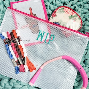 May include: A collection of craft supplies, including embroidery floss in various colors, a clear zippered pouch with "WIP" embroidered on it, a small floral print pouch, and a pink flexible neck light, all arranged on a textured teal surface.
