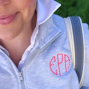 May include: A light gray zip-up sweatshirt featuring a white collared shirt underneath. The sweatshirt has a circular monogram embroidered in bright pink thread on the left chest with the letters EPA. A gray and white strap rests on the shoulder.