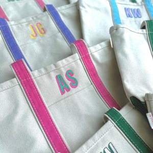 May include: A collection of canvas tote bags with colorful accents. Each bag features a different set of embroidered initials in vibrant colors. The handles and trim come in various shades, including pink, blue, and green, creating a personalized and stylish look.