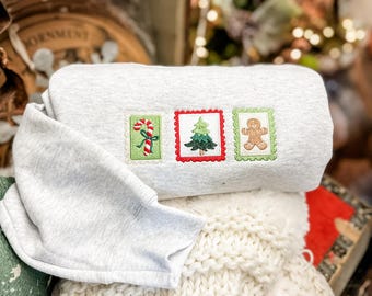 Könnte beinhalten: Ein hellgraues Sweatshirt mit gestickten Weihnachts-Briefmarkenmotiven. Die Briefmarken zeigen eine Zuckerstange, einen Weihnachtsbaum und einen Lebkuchenmann. Das Sweatshirt liegt gefaltet auf einer cremefarbenen Strickdecke.
