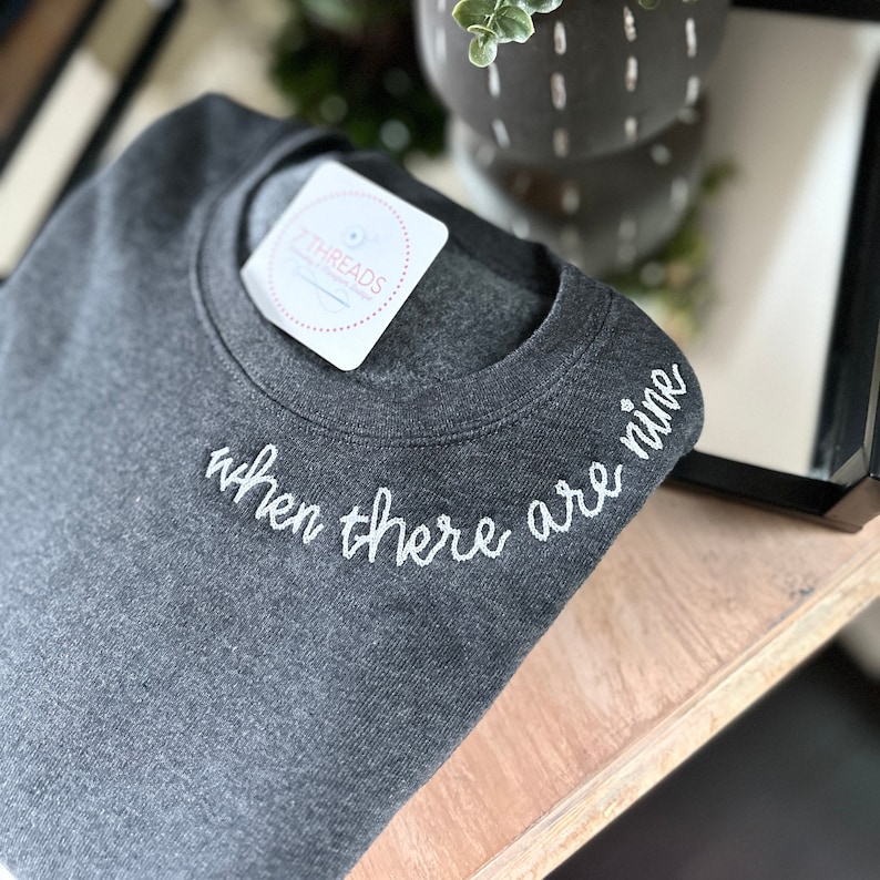 When There Are Nine chain stitch embroidery in white thread, along collar of dark gray comfortable sweatshirt