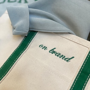 May include: A cream-coloured canvas tote bag with dark green vertical stripes and the text "on brand" embroidered in dark green script. A light blue folded garment with the partial text "UCKY" is resting on top of the tote bag.