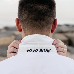 May include: A white button-down shirt with the date "10-10-2026" embroidered in black on the collar. The image shows the back of a person wearing the shirt, with hands gently touching the collar.