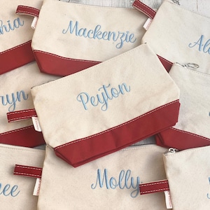 Peut inclure: Sacs à cosmétiques en toile crème avec des bases et des poignées rouges. Chaque sac est brodé d'un nom différent en écriture bleu clair. Les sacs sont zippés et semblent personnalisés.