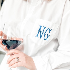 Peut inclure: Chemise blanche boutonnée avec un monogramme brodé bleu sur la poche. La chemise est à manches longues et possède un col classique. Un verre de vin rouge est tenu à la main.