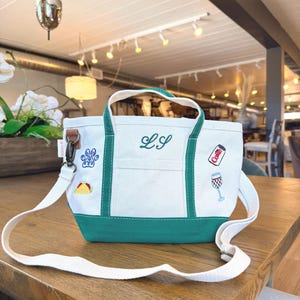 May include: White and teal tote bag with a shoulder strap. The bag has embroidered patches of a paw print, a taco, a Coke can, and a wine glass. The initials "LS" are embroidered on the front.