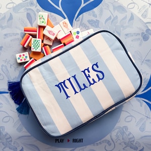 Könnte beinhalten: Ein blau-weiß gestreiftes Etui mit dem Wort „TILES“ in blauen Buchstaben, umgeben von bunten Mahjong-Steinen. Das Etui hat eine marineblaue Umrandung und eine blaue Quaste. Der Hintergrund zeigt ein blaues Blumenmuster.