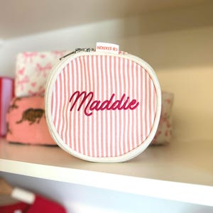 May include: A round, zippered pouch with pink and white vertical stripes. The name "Maddie" is embroidered in cursive script. The pouch has a white border and a silver zipper. The text "CB STATION" is printed on a small tag.