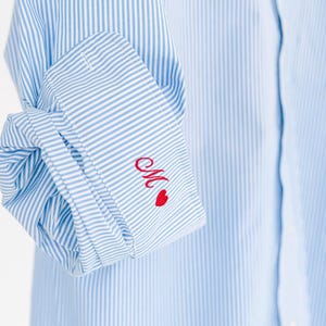 May include: A light blue and white pinstriped long-sleeve shirt with a folded cuff. The cuff features red embroidered cursive lettering of the letter M followed by a small red heart symbol, adding a distinct detail to the classic striped fabric.