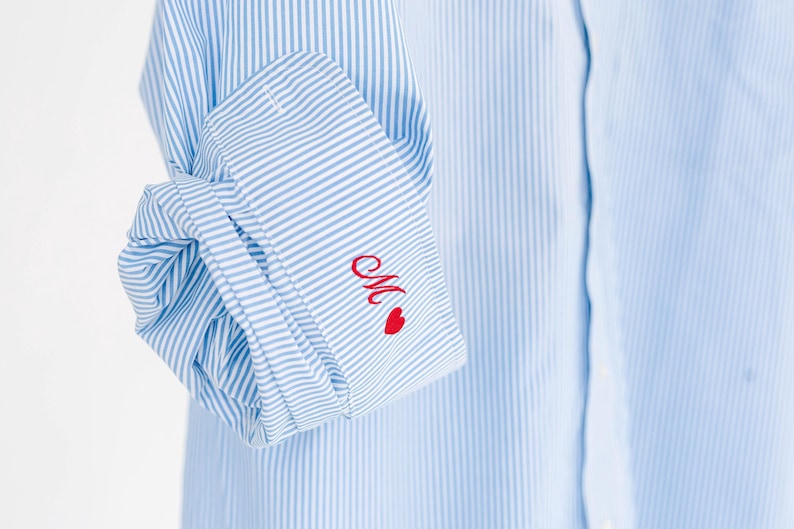 May include: A light blue and white pinstriped long-sleeve shirt with a folded cuff. The cuff features red embroidered cursive lettering of the letter M followed by a small red heart symbol, adding a distinct detail to the classic striped fabric.