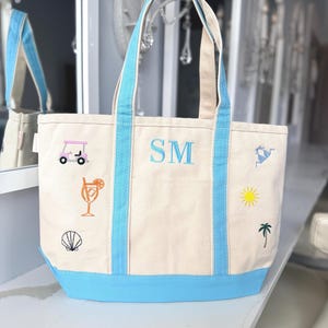 Könnte beinhalten: Eine beigefarbene Tragetasche aus Segeltuch mit hellblauen Rändern und Griffen. Die Tasche ist mit gestickten Symbolen versehen, darunter ein rosa Golfwagen, ein orangefarbener Cocktail, eine Muschel, ein blauer Bikini, eine gelbe Sonne und eine grüne Palme, sowie den blauen Buchstaben SM.