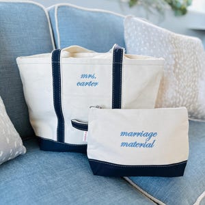 May include: A cream-colored canvas tote bag and a matching zippered pouch with navy blue trim. The tote features the embroidered text "mrs. carter" in blue, while the pouch displays the embroidered text "marriage material" in blue.