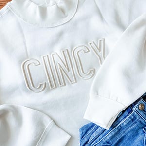 May include: A white sweatshirt with the word "Cincy" embroidered in a light beige color. The sweatshirt has a mock neck and long sleeves. Blue denim jeans are visible in the bottom right corner.