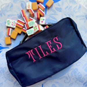 May include: A navy blue zippered pouch embroidered with the word "TILES" in pink, overflowing with colorful mahjong tiles. The tiles are orange, white, and green, scattered on a blue and white patterned surface. The pouch is rectangular and appears to be made of a smooth fabric.