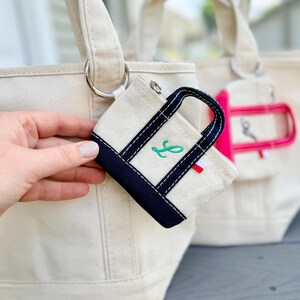May include: A miniature off-white canvas pouch with navy blue trim and handles, featuring a green embroidered script letter. The pouch includes a small silver zipper and a tiny red, white, and blue tag, attached to a larger matching canvas tote bag.