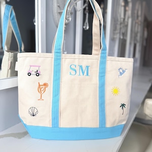 May include: Cream-colored canvas tote bag with light blue accents. Features embroidered beach-themed designs, including a golf cart, cocktail, seashell, sun, palm tree, and sailboat. The initials "SM" are embroidered in blue.