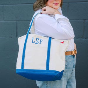 May include: A cream canvas tote bag with blue trim and handles. The bag has the embroidered initials "LSP" in blue. The base and handles are blue. A small red tag is visible on the side. The bag is a practical accessory.
