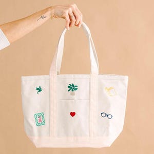 May include: A cream-colored canvas tote bag featuring embroidered icons including a green plant in a pot, a red heart, blue glasses, a green dragon, a yellow book, and a green rectangular frame with a pink flower design on the front panel.