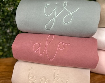 Può includere: Una pila di tre felpe piegate in verde salvia, rosa antico e beige. Ogni felpa presenta un ricamo in corsivo personalizzato sul davanti: quella in alto riporta ejs, quella centrale alo e quella in basso kmb con fili in colori coordinati.