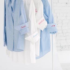 May include: Four button-down shirts hanging on a rack. The first is light blue with 'Sarah' embroidered on the cuff. The second is white with 'Bride' on the cuff. The third is white with '04.10.27' on the cuff. The fourth is blue striped with 'M' and a heart.