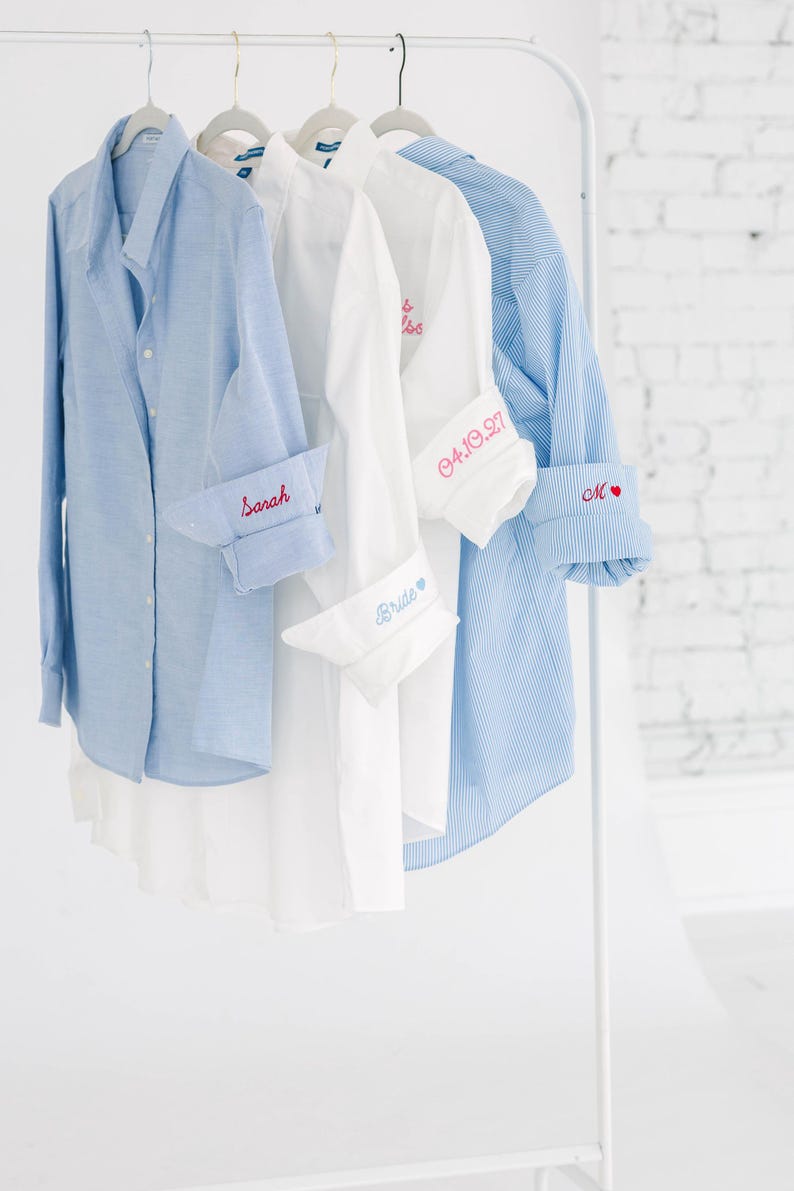 May include: Four button-down shirts hanging on a rack. The first is light blue with 'Sarah' embroidered on the cuff. The second is white with 'Bride' on the cuff. The third is white with '04.10.27' on the cuff. The fourth is blue striped with 'M' and a heart.