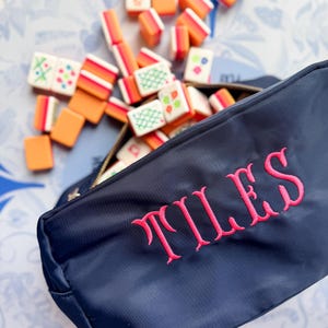May include: Navy blue zippered pouch embroidered with the word "TILES" in pink. The pouch is open, revealing a collection of colorful Mahjong tiles. The tiles are orange, white, and green, scattered around the pouch.