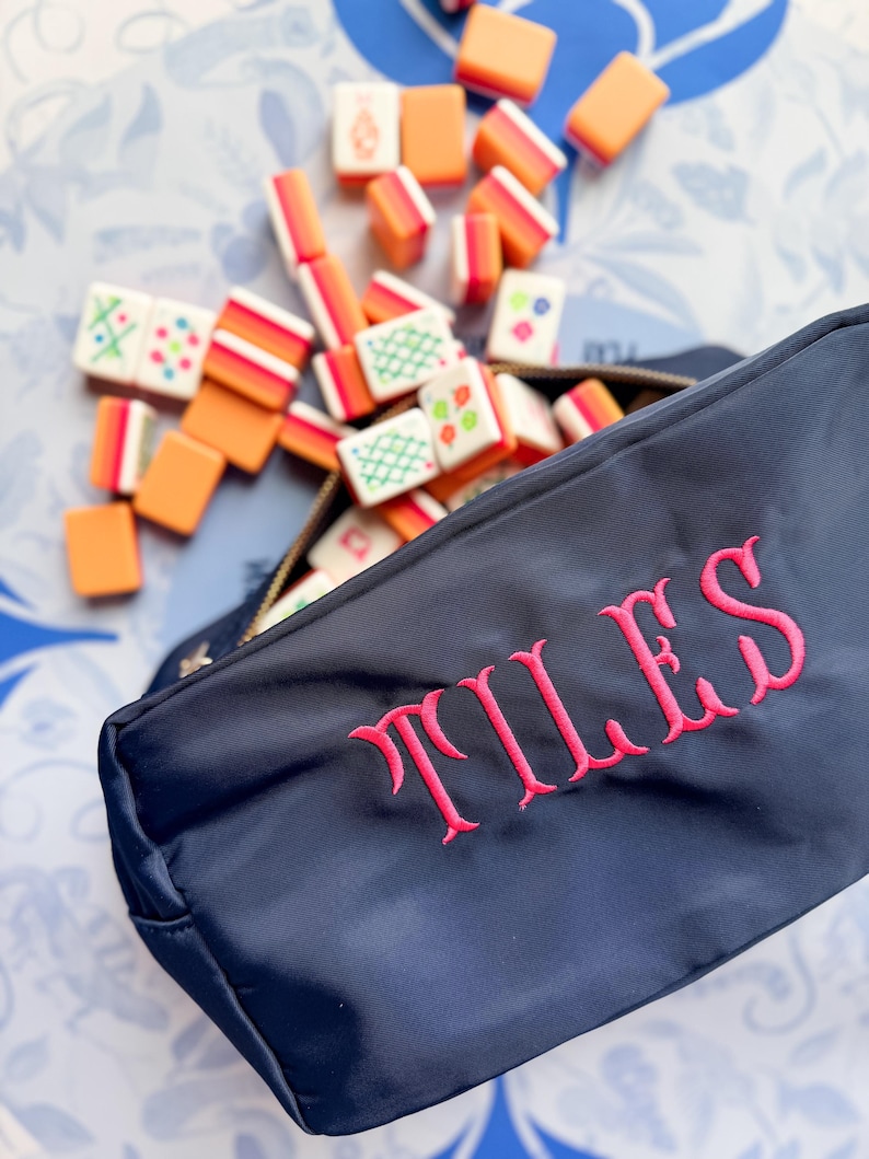 May include: Navy blue zippered pouch embroidered with the word "TILES" in pink. The pouch is open, revealing a collection of colorful Mahjong tiles. The tiles are orange, white, and green, scattered around the pouch.