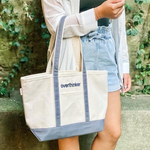 May include: A cream-colored canvas tote bag with a reinforced blue base and matching blue vertical straps. The word "overthinker" is embroidered in blue lowercase letters on the front center of the bag, which features an open top design.