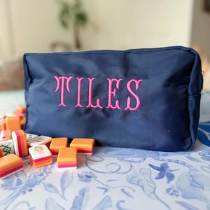 May include: A navy blue zippered pouch with the embroidered word "TILES" in pink. The pouch is surrounded by colorful mahjong tiles on a patterned blue and white surface. The image suggests a game-related theme.