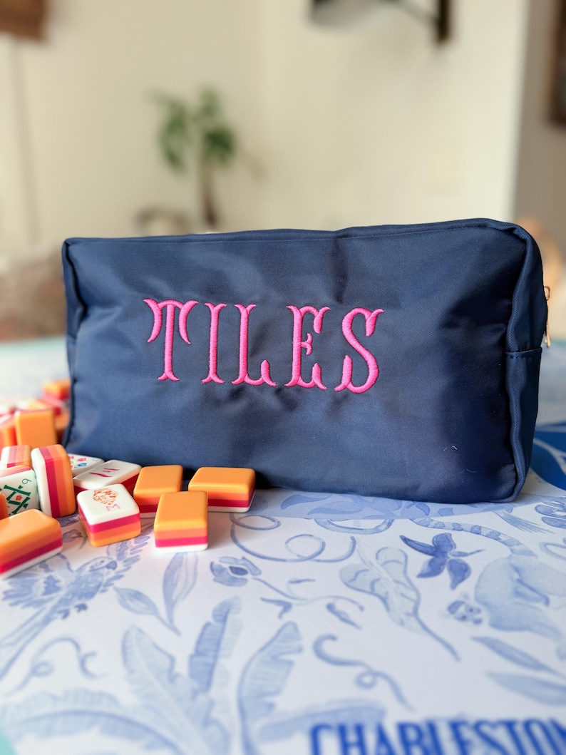 May include: A navy blue zippered pouch with the embroidered word "TILES" in pink. The pouch is surrounded by colorful mahjong tiles on a patterned blue and white surface. The image suggests a game-related theme.